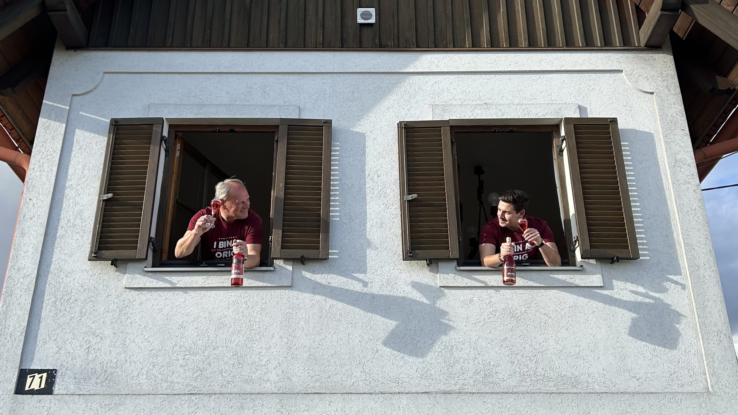 Uhudler Berzkovics Vater und Sohn schauen aus zwei Fenstern im Haus und grüßen einander zu.